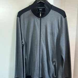 Grå zipjacka från BOSS - Snygg grå zipjacka från BOSS med svarta axelpartier och diskret logga på bröstet. Jackan har hel dragkedja framtill, två fickor med dragkedja och hög krage. Perfekt för en clean och sportig look. nyskick