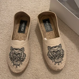 Beige espadrillos från Kenzo med tiger - Snygga beige espadrillos från Kenzo med ikoniskt broderat tigermotiv framtill. Skorna har en klassisk rund tå och flätad sula i rep. Ovandelen är i bomull och insidan har en naturlig look. Perfekta för en chill och trendig sommarstil.