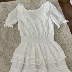 Vit volangklänning från Gina Tricot - Superfin vit kortklänning från Gina Tricot med volanger, smock i midjan och korta puffärmar. Klänningen har omlottkänsla i ryggen med knyt och är gjord i ett lätt, luftigt material. Perfekt för sommarens alla tillfällen!