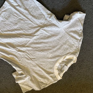 Grå Adidas t-shirt med logga - Snygg grå t-shirt från Adidas med klassisk vit Adidas-logga broderad på bröstet. T-shirten har rund hals med vit ribbad kant och är kortärmad. Perfekt för dig som gillar enkel och sportig stil. Materialet är mjuk bomull som känns skönt mot huden. KÄNNS INTE som en Large skulle nog mer säga S