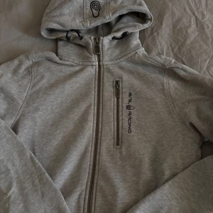 Grå hoodiejacka från Sail Racing - Snygg grå hoodiejacka från Sail Racing med hel dragkedja, bröstficka med dragkedja och broderad logga på bröstet. 