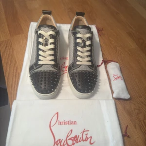 Christian Louboutin sneakers med nitar - Snygga sneakers från Christian Louboutin med signifikant röd sula och coola nitar på tån. Skorna har svart och grå textil med svarta skinnpaneler och vita kontrastsömmar. Klassisk snörning och rund tå. Perfekt för dig som vill sticka ut med exklusiv stil. Nypris ca 10 000, helt nya aldrig använda. 