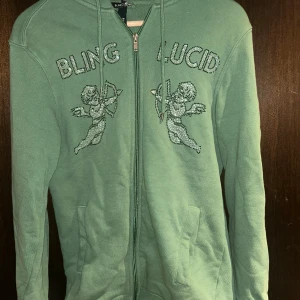 Grön hoodie från Blinglucid med rhinestones - Snygg grön hoodie från Blinglucid med dragkedja, huva och snörning. Framsidan har coola strassdetaljer med texten 'BLING LUCID' och två änglar. Perfekt för dig som gillar streetstyle och vill sticka ut lite extra.