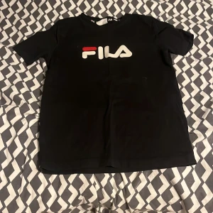 Svart FILA t-shirt med logga - Svart t-shirt från FILA med stor, vit och röd logga på bröstet. Klassisk rund hals och korta ärmar. Skön passform och tillverkad i mjuk bomull, perfekt för chill dagar. Passar dig som gillar sportiga och stilrena plagg. Ordinarie pris:450kr