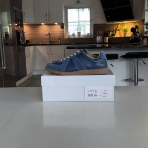Blå Maison Margiela sneakers i skinn och mocka - Maison Margiela sneakers i blått skinn och mocka med beige snörning och innersida. Snygg kontrast mot den bruna gummisulan. Klassisk siluett med rund tå och platt sula, perfekt för en clean och stilren look.