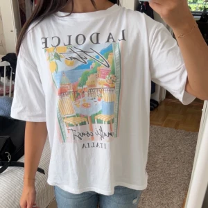 Vit t-shirt med tryck från Chica London - Vit t-shirt från Chica London med färgglatt tryck framtill som visar en italiensk kuststad och texten 'La Dolce Vita'. Aldrig använd! Modellen är oversize, och i onesize, nypris 300!