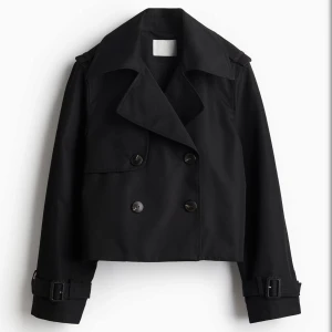 Trenchcoat kort - Snygg svart croppad trenchcoat med bred krage, dubbelknäppning och bältdetaljer vid ärmsluten. Jackan har en loose passform och stora knappar framtill. Perfekt för dig som gillar en stilren och trendig look.