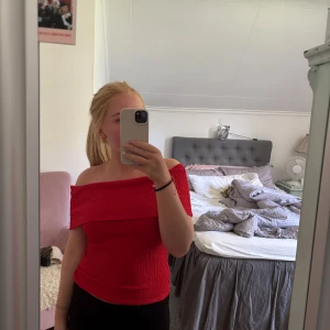 Röd offshoulder topp med struktur - Säljer en snygg röd offshoulder topp med smockad struktur och croppad passform. Toppen är ärmlös och har en bred volangkant över axlarna som ger en trendig look. Perfekt att styla med jeans eller kjol för en cool vibe.