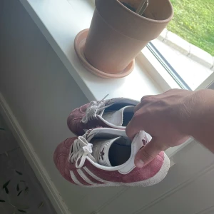 Adidas Gazelle i vinröd mocka - Säljer ett par Adidas Gazelle sneakers i vinröd mocka med vita detaljer och klassiska tre ränder på sidorna. Skorna har vit plös och snörning samt rund tå. Perfekta för dig som gillar retrostil och vill sticka ut lite extra.