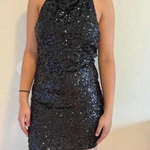 Svart glittrig paljettklänning - Säljer en svart kort paljettklänning med halterneck och öppen rygg. Klänningen är täckt av glittriga paljetter som ger en riktigt festlig vibe. Tight passform som framhäver figuren och den är ärmlös för en snygg look. Perfekt för dig som vill sticka ut på festen!😀😍