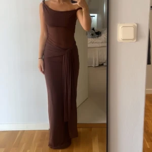 Brun långklänning med omlott och drapering - Säljer en snygg brun långklänning med omlott-detalj och drapering vid midjan. Klänningen har en asymmetrisk axelbandslös design och sitter tight mot kroppen för en smickrande siluett. Perfekt för dig som gillar stilrena och trendiga plagg.