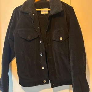 Svart manchesterjacka från Vailent - Svart manchesterjacka från Vailent med mysigt teddyfoder och krage. Klassisk jeansjacke-modell med två bröstfickor med lock och knappar framtill. Perfekt för dig som gillar en stilren och tidlös look med lite extra värme. Väldigt bra skick. Passar perfekt