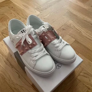Valentino garvani skor - Säljer ett par Valentino Garvani sneakers i vitt skinn med rosa band och ikoniska pyramidnitar. Snygg och chunky sula med mönster, rund tå och klassisk snörning. 