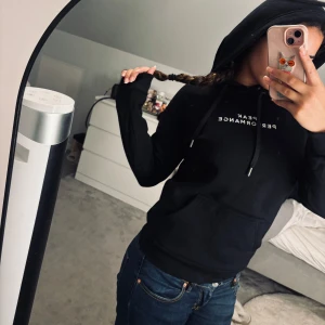 Svart hoodie från Peak Performance - Svart hoodie från Peak Performance med vit logga på bröstet. Klassisk modell med huva och snörning, samt stor magficka framtill. Mjuk och skön bomullskvalitet, perfekt för chill eller träning. Snygg passform och ribbade muddar vid ärmslut och nederkant.