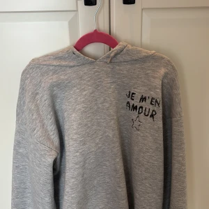 Grå hoodie med tryck och huva - Säljer en grå hoodie med coolt svart tryck 'JE M'EN AMOUR' och en stjärna på både fram- och baksidan. Tröjan har en stor huva, långa ärmar och ribbade muddar. Perfekt för chill dagar och streetstyle. Materialet känns mjukt och skönt mot huden.