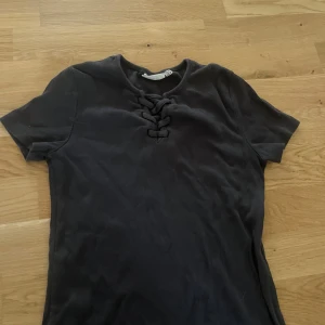 Svart ribbad topp med snörning Kappahl - Svart ribbad topp från Kappahl med korta ärmar och snygg snörningsdetalj vid halsringningen. Toppen har en tight passform och är gjord i mjukt, stretchigt material som sitter skönt på kroppen. Perfekt för dig som gillar stilrena basplagg med en twist.