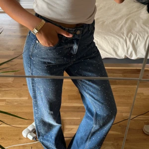 Blå jeans med strassdetaljer - Säljer ett par blå jeans med hög midja och raka ben, täckta av små glittriga strassdetaljer över hela byxorna. Jeansen har klassisk femficksmodell och är tillverkade i kraftigt denimtyg. Perfekta för dig som vill sticka ut lite extra.