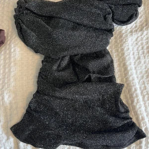 Svart glittrig offshoulder topp NA-KD - Supersnygg svart topp från NA-KD med glittrigt tyg och offshoulder-design. Toppen har en tight passform och är perfekt för dig som vill sticka ut med lite extra glitter. Materialet är mjukt och följsamt, och axelpartiet har en snygg drapering.