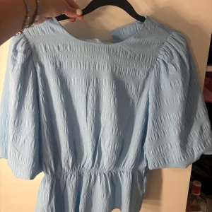 Ljusblå blus från Studio, storlek S - Supersöt ljusblå blus från Studio med puffiga korta ärmar och smockad struktur. Blusen har rund halsringning och markerad midja som ger en snygg siluett. Perfekt för dig som gillar en feminin och trendig look.