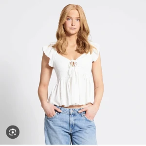Vit blus med volangärm och knyt - Supersöt vit blus med volangärmar och v-ringning. Toppen har knyt fram och en luftig, flowy passform som ger en romantisk vibe. Perfekt till jeans eller kjol för en fräsch look. Materialet känns lätt och somrigt.