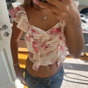 Blommig croppad blus med volang - Supersöt blommig croppad blus med volangdetaljer och v-ringning. Toppen har ljusa och rosa toner, är ärmlös med breda volangaxlar och smickrande rynk framtill. Perfekt för varma dagar och somriga vibes. Bilden är lånad från förra ägaren❤️
