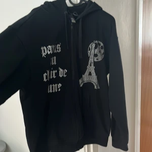 Svart hoodie med Paris-tryck - Svart hoodie med dragkedja och huva med detaljerad text och bild med rinestones. Fungerar i alla tillfällen. Storlek S men passar även M. Väldigt bra skick