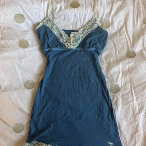 Abercrombie babydoll linne - Supersöt blå kortklänning från Abercrombie & Fitch med krämvit spets vid v-ringad hals och nederkant. Axelbandslös modell med stretchigt material som sitter snyggt. Liten vit logga broderad längst ner. Perfekt för sommarens alla tillfällen!