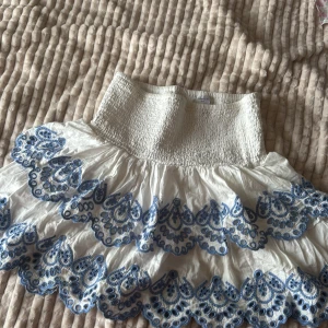 Vit kjol med blå broderi från Lindex - Supersöt vit kortkjol från Lindex med bred smockad midja och två lager volanger. Kjolen har detaljerad blå broderi längs volangerna som ger en fräsch och somrig vibe. Perfekt för dig som gillar unika detaljer och vill sticka ut lite extra.
