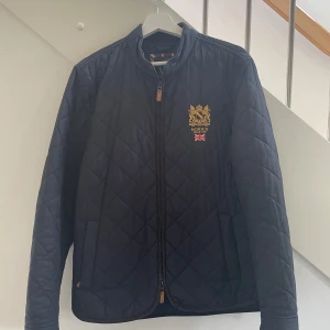 Quiltad jacka från Morris, marinblå M - Snygg quiltad jacka från Morris i marinblått med guldbroderad logga och brittisk flagga på bröstet. Jackan har dragkedja framtill, ståkrage och klassiskt rutmönster. Perfekt för dig som vill ha en stilren jacka nu under hösten. |Tveka inte att skriva vid funderingar ☺️|