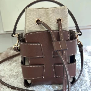 Brun bucket bag från H&M - Snygg bucket bag från H&M i brun skinnimitation med kontrastsömmar och beige canvasinsida. Väskan har både handtag och axelrem samt cool snörning upptill. Perfekt för dig som vill ha en trendig och praktisk accessoar med unik design.