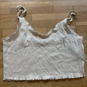 Beige ribbad croppad linnetopp - Supersöt beige croppad linnetopp från Only med tunna axelband och ribbad struktur. Toppen har små hålmönster i tyget och vågig kant vid halsringning och nederkant. Perfekt för varma dagar och enkel att matcha med jeans eller kjol.