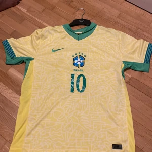 Brasilien Brazil  Nike fotbollströja - Säljer en Brasilien fotbollströja med Rodrygo och nummer 10 på ryggen. Tröjan är gul med gröna detaljer, har ett subtilt grafiskt mönster och grönt Nike-märke samt CBF-emblem på bröstet. Kortärmad och tillverkad i lätt polyester som andas.