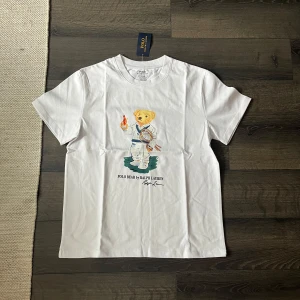 Ralph Lauren T-shirt  - Helt ny Vit t-shirt från Polo Ralph Lauren med klassisk Polo Bear-tryck på framsidan där björnen håller en fackla och tennisracket. Rund hals, korta ärmar och slim fit. Materialet är mjuk bomull och passformen är modern och snygg  .