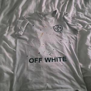 Vit Off-White t-shirt med tryck - Säljer en vit t-shirt från Off-White med coolt tryck fram och bak. Framsidan har färgstänk och logga, baksidan har grafiskt motiv med text och banddetaljer. T-shirten är kortärmad och har en relaxed fit. Perfekt för dig som gillar streetwear och statement-plagg.