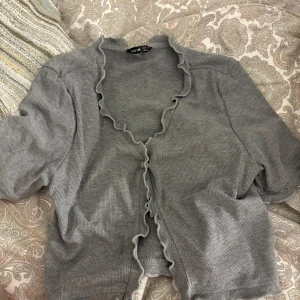 Grå croppad bolerotopp från SHEIN - Grå bolerotopp från SHEIN med croppad passform och volangkant längs öppningen. Toppen är ribbad och har korta ärmar, perfekt för att ge din outfit en chill vibe. Enkel att matcha med jeans eller kjol för en snygg look.