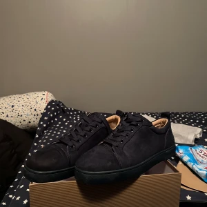 Svarta mockasneakers från Louboutin - Snygga svarta sneakers i mocka från Christian Louboutin med klassiska röda sulor. Skorna har rund tå, svarta snören och diskret design som ger en lyxig vibe. Perfekta för dig som vill sticka ut med exklusiva detaljer och stilren look.