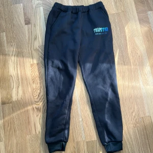 Svarta mjukisbyxor Trapstar - Svarta mjukisbyxor från Trapstar med tryck i blått och vitt på ena benet. Byxorna har resår i midjan, muddar vid bensluten och två sidofickor. Perfekta för chill dagar eller streetwear-stil.