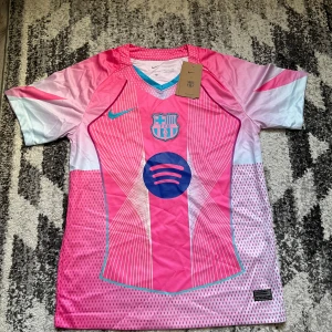  Barcelona tröja rosa - Snygg FC Barcelona fotbollströja från Nike i rosa och ljusblått med vita detaljer. Tröjan har korta ärmar, klubbmärke på bröstet och Spotify-logga i mitten. Tillverkad i lätt och ventilerande material, perfekt för träning eller match.