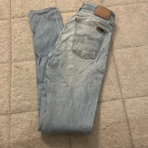 Ljusblå Slim Nudie jeans - Storlek: W29, L34. Säljer ett par ljusblå Nudie jeans med klassisk femficksdesign och läderpatch bak i midjan. Jeansen har en rak passform och är tillverkade i mjukt denimtyg. Perfekta för dig som gillar en avslappnad men ändå stilren look.