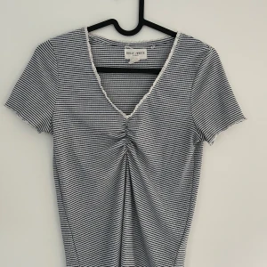 Randig topp med v-ringning Lindex - Superfin kortärmad topp från Lindex med smala svarta och vita ränder, v-ringning med spetskant och snygg rynkdetalj framtill. Mjuk ribbad och figurnära passform. Perfekt till jeans eller kjol för en gullig look.