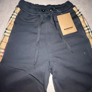 Svarta Burberry mjukisbyxor med rutor - Svarta mjukisbyxor från Burberry med klassiska rutiga paneler längs sidorna. Byxorna har resår i midjan med snörning och är tillverkade i mjukt bomullsmaterial. Perfekta för en chill och stilren look.