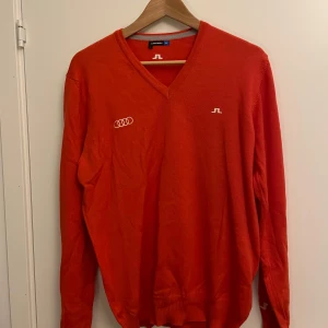 Orange stickad tröja från J.Lindeberg - Snygg orange stickad tröja från J.Lindeberg med vit logga på bröstet och ryggen samt Audi-märke på framsidan. Tröjan har v-ringad hals och långa ärmar, perfekt för dig som gillar stilren design och sportiga detaljer. Materialet är merinoull.