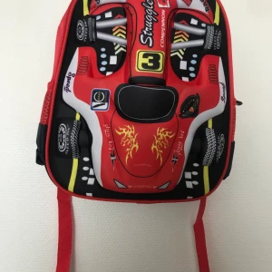 Röd ryggsäck med bilmotiv - Cool ryggsäck i form av en sportbil med röda, svarta och gula detaljer. Framsidan har 3D-effekt med tryck av däck, eldflammor och racingnummer. Justerbara röda axelband och dragkedja. Perfekt för dig som gillar bilar och vill sticka ut i skolan. Helt ny 