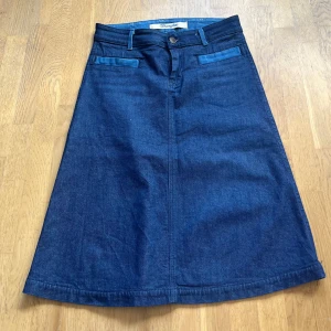 Wrangler lång jeanskjol blå W29 - Säljer en snygg lång jeanskjol från Wrangler i mörkblå denim. Kjolen har klassiska fickor fram och bak, bälteshällor och knappstängning. Den har en rak, lätt utsvängd form och är tillverkad i bomull. Perfekt för dig som gillar retrovibe och denim.