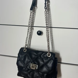 Svart axelväska med kedja - Snygg svart axelväska i quiltat skinnimitation med silverfärgad kedjerem och vridlås. Väskan har ett rymligt fack och en innerficka med dragkedja. Perfekt storlek för det viktigaste och ger en trendig vibe till din outfit.