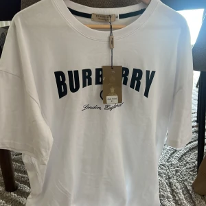 Vit Burberry t-shirt med logga - Vit t-shirt från Burberry med svart tryckt logga på bröstet och texten 'London, England' under. Klassisk rund halsringning och korta ärmar. Tillverkad i mjuk bomull för en clean och stilren look.