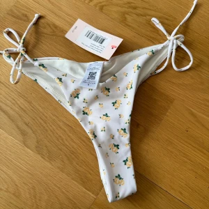 bikiniunderdel - Supersöt bikiniunderdel från Nelly i vitt med små gula blommor. Modellen har knyt i sidorna och en scrunch-detalj bak. Helt ny