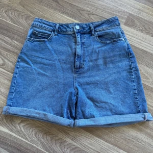 Blåa jeansshorts från Vila, hög midja - Snygga blå jeansshorts från Vila med hög midja och klassisk femficksdesign. Shortsen har uppvikta benslut och bälteshällor, perfekta för en chill sommarlook. 