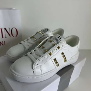 Valentino Rockstud vita sneakers - Snygga vita Valentino Rockstud sneakers i skinn med ikoniska guldiga nitar och coola detaljer på sidorna och hälen. Klassisk rund tå, platt sula och snörning framtill. Perfekta för dig som vill sticka ut med en lyxig touch.