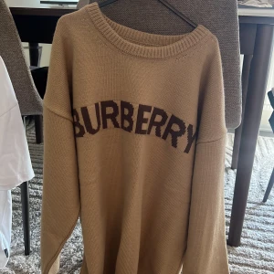 Beige stickad tröja från Burberry - Snygg beige stickad tröja från Burberry med brunt logotyp-tryck framtill och stor B-detalj på ryggen. Tröjan har rund halsringning och långa ärmar, perfekt för dig som gillar klassisk men trendig stil. 